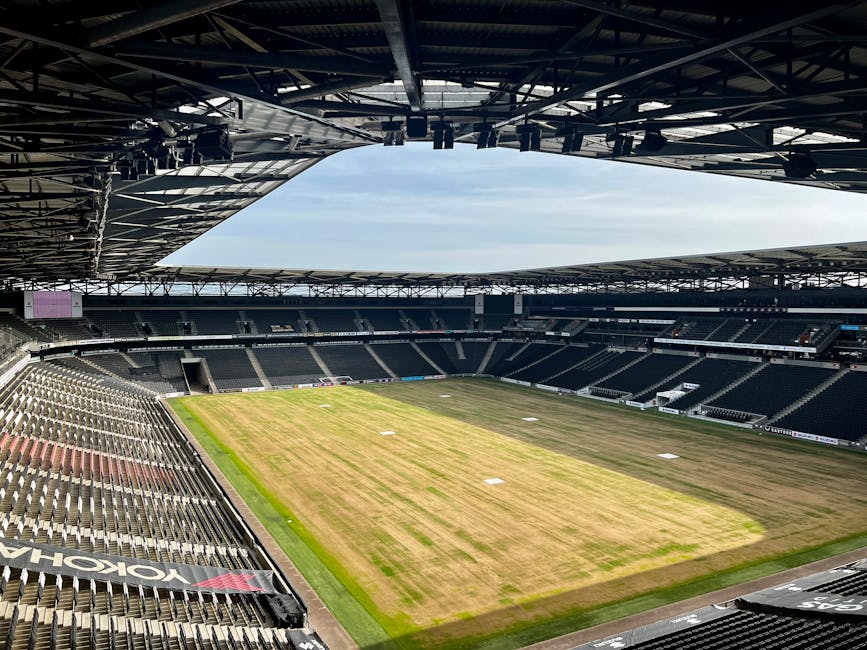 The image shows a panoramic view of an empty sports stadium, likely a cricket or football ground, with a partially covered roof structure overhead and rows of dark-colored seating surrounding the pitch area. The playing field in the center is grassed but appears to be in the process of being cleaned or prepared, with patches of dirt or dried grass visible. The stadium's seating sections are predominantly black or dark grey, extending in curved lines around the pitch, with some empty chairs visible along the front and sides. The environment is well-lit with natural daylight, and the sky above is partly cloudy. The surrounding environment suggests an indoor or semi-enclosed space designed for large-scale sporting events. This scene may relate to private or independent event preparation or ground maintenance, which can involve waste removal and site clearance, typical services offered by companies such as Rubbish Removal Twickenham in the context of stadium or venue event management, ensuring the space is cleared of debris and waste after use or during preparation for upcoming events.