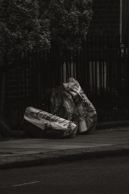 Two large black plastic rubbish bags, appear to be filled with waste, sitting on the pavement close to a street curb. The bags are slightly crumpled and have a glossy finish, indicating they are made of durable plastic. One bag is partially leaning against a black metal fence with vertical bars, which separates the sidewalk from a residential property. Behind the fence, there are trees with dense foliage, and the scene is shaded, suggesting it is either early morning or late afternoon. The pavement surface shows signs of regular use, with a visible curb edge and a section of road in the foreground. The scene overall suggests a typical urban or suburban environment where private rubbish collection might be arranged by a local waste management service such as Rubbish Removal Twickenham, demonstrating an example of on-site clearance or alternative waste handling outside of council waste collection areas.