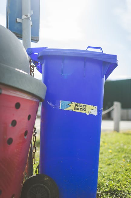 A large, upright blue plastic rubbish bin with a closed lid, positioned on a paved surface near a grassy area. The bin has a yellow and blue sticker that reads 'FIGHT BACK!' adhered vertically on its front. To the left of the blue bin, part of a pink rubbish bin with small black holes in its side is visible, along with a portion of its lid. The scene is outdoors with natural daylight, showing a clear sky with some clouds, a metal pole supporting a sign or structure in the background, and a guardrail at a distance. The environment appears to be a residential or public area, and the image emphasizes waste collection and disposal options, which may relate to private rubbish removal services such as those offered by Rubbish Removal Twickenham, featured on the page about alternative waste handling methods.